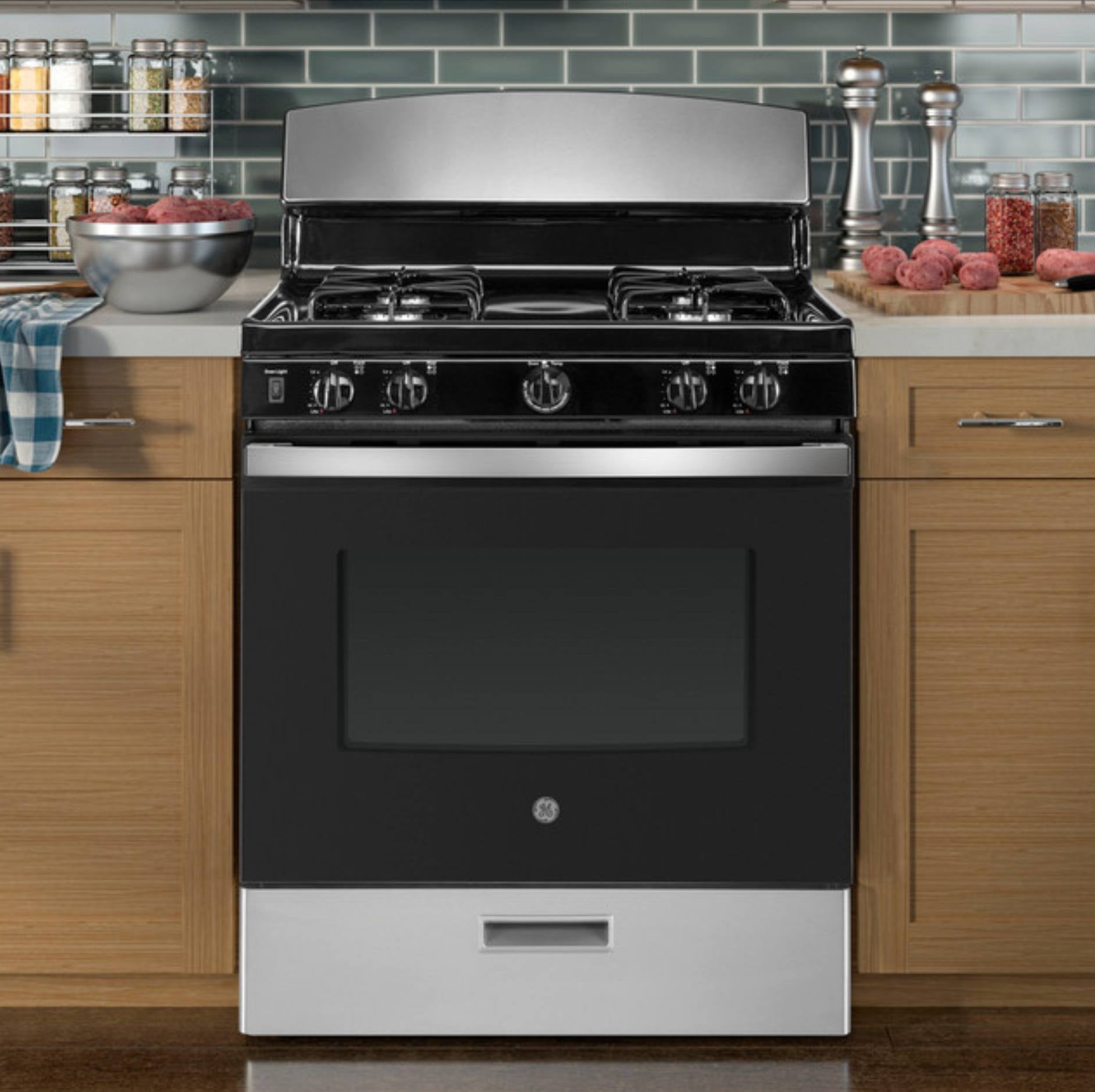 A freestanding gas range stove with a black oven door, stainless steel control panel and backguard, set in a kitchen with wood cabinets and a tiled backsplash.