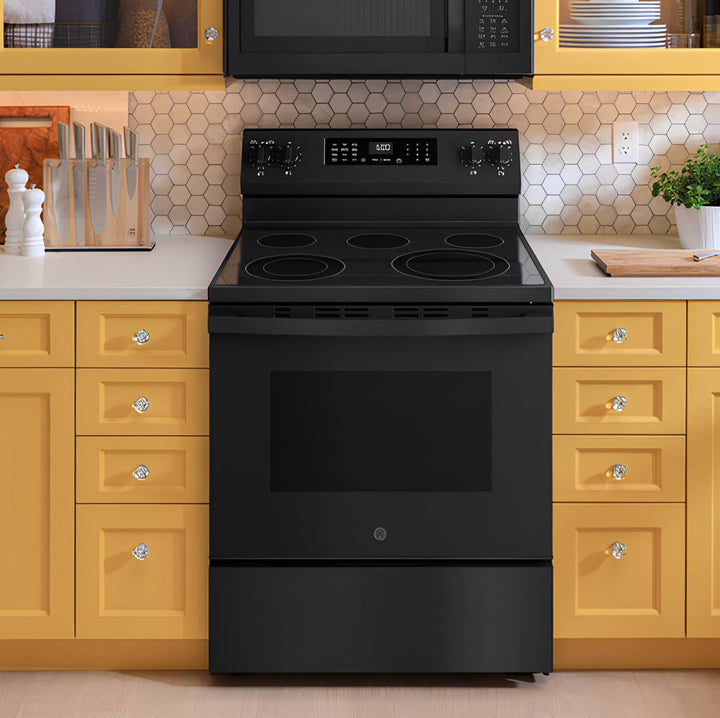 A GE freestanding electric range in black, set in a kitchen with yellow cabinets, white countertops, and a hexagonal tile backsplash.
