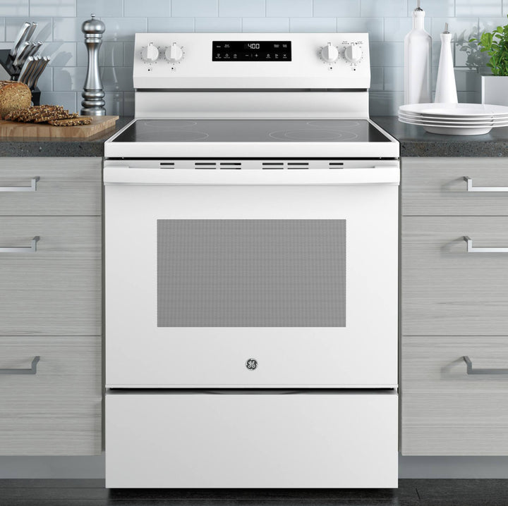 A white GE freestanding electric range with a glass cooktop, digital control panel, and lower storage drawer, set against a modern kitchen background with gray cabinets and light blue tile.