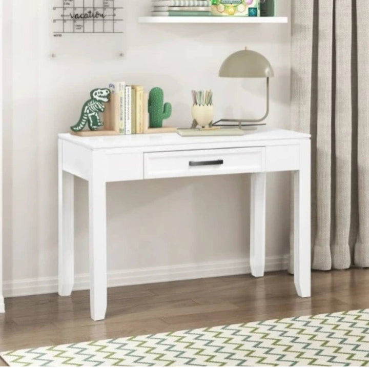 A white wooden writing desk with a single drawer and black metal handle, styled in a home office setting with a lamp, books, and a dinosaur figurine on top.