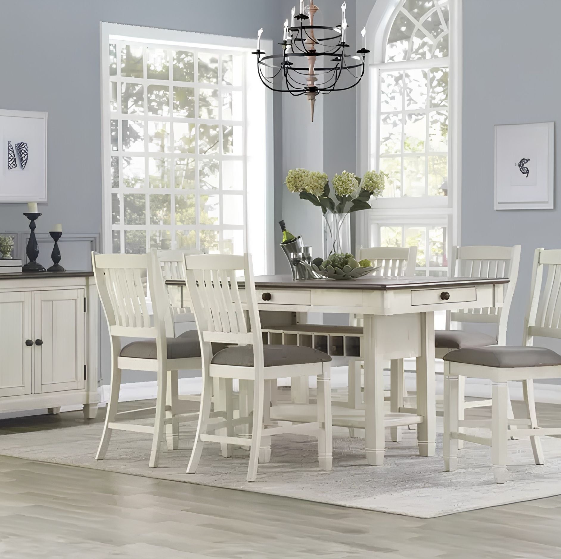 A white wooden dining table set with six matching high-back chairs with gray upholstered seats, placed in a bright dining room with light gray walls, large windows, and a chandelier overhead.