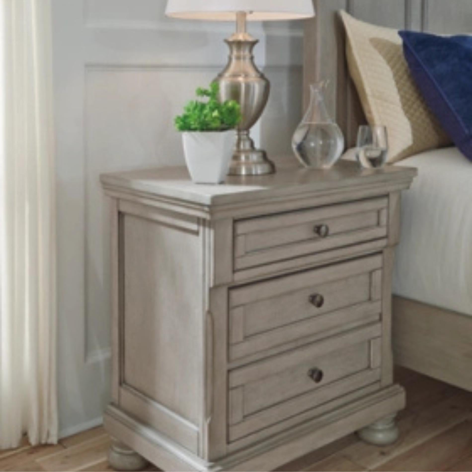 Wooden nightstand with drawers, lamp, and decorative items in a bedroom setting.
