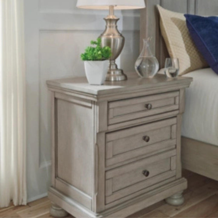 Wooden nightstand with drawers, lamp, and decorative items in a bedroom setting.