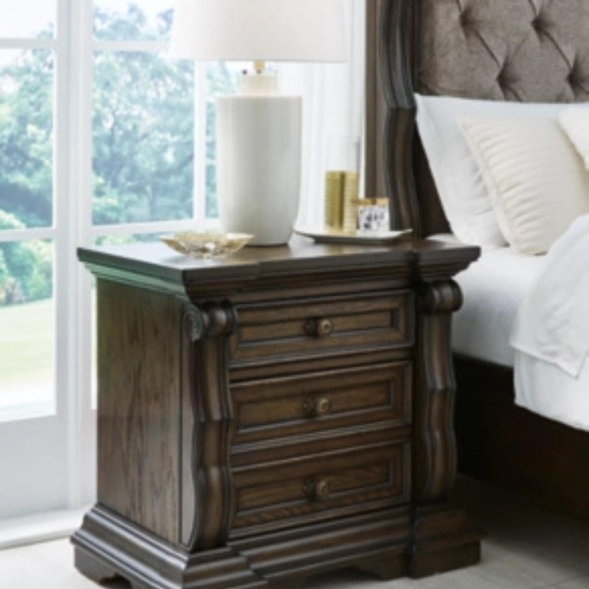 Wooden nightstand with lamp and decor items next to a bed with white bedding.