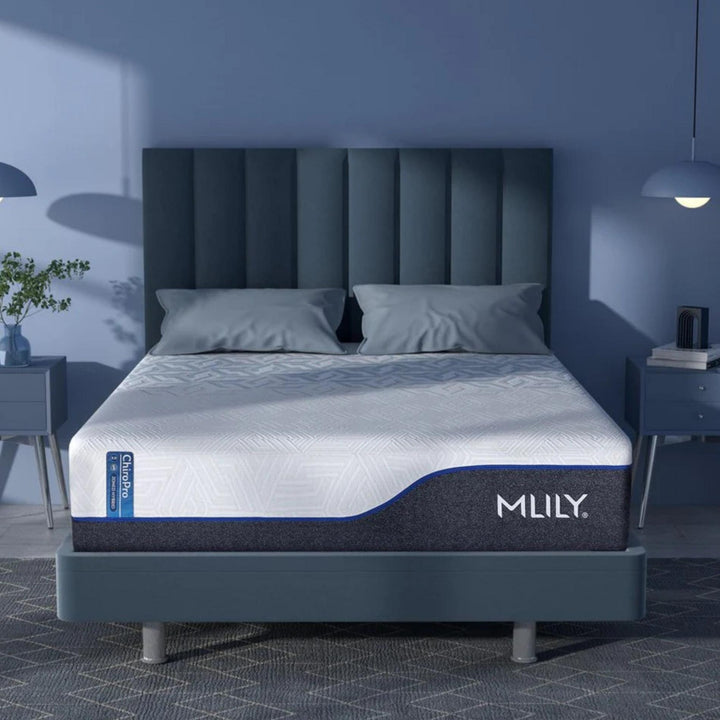 An MLILY mattress shown on a matching bed frame in a modern bedroom setting. The bed features a dark slate blue headboard and nightstands, with soft blue ambient lighting casting shadows on the walls.