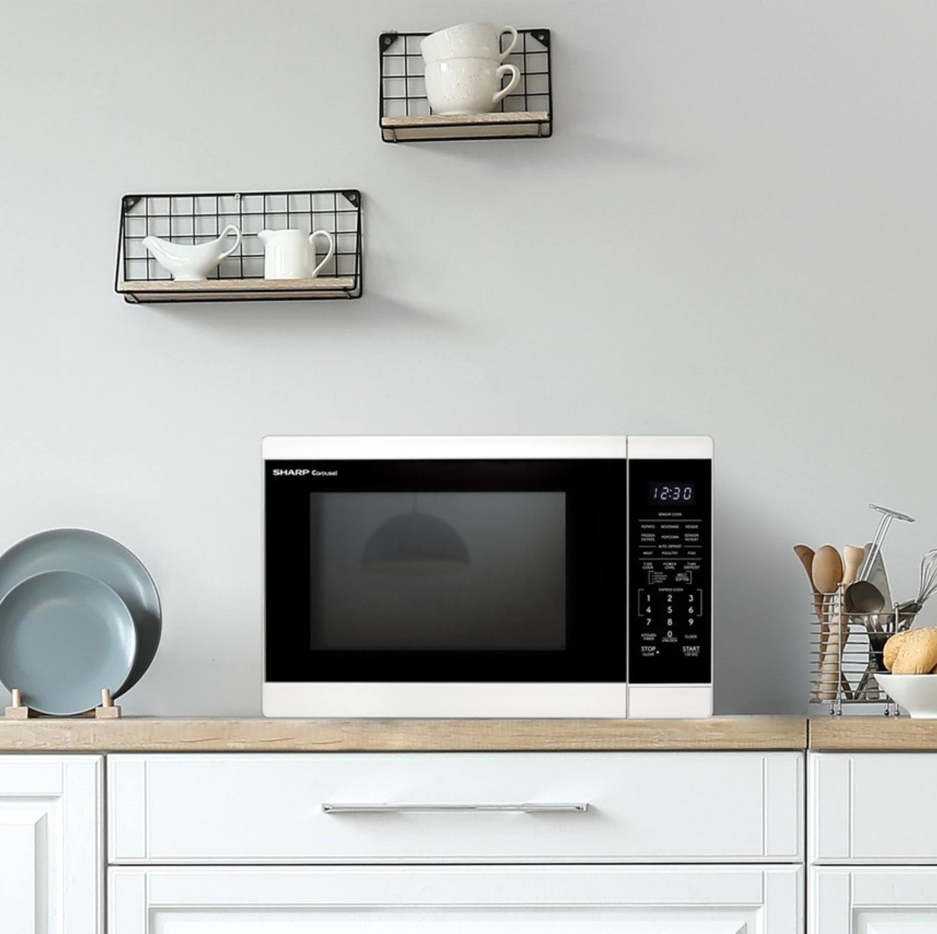 A Sharp microwave oven sits on a light wood kitchen countertop against a white wall, with two black wire wall shelves above holding white mugs and a pitcher. White cabinets are visible below.