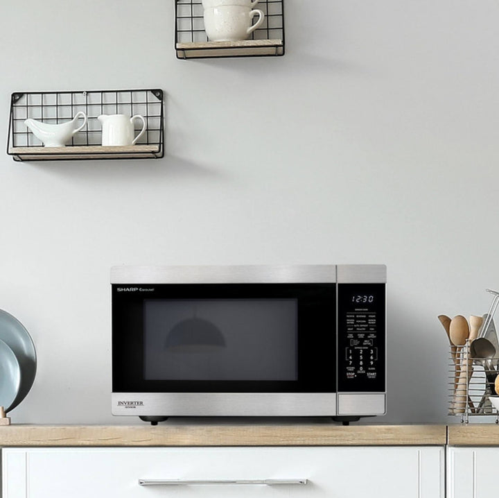 A countertop microwave oven with a stainless steel exterior and black glass door, placed on a light wood cabinet in a kitchen setting. Two black wire wall shelves with white dishes are mounted above, and a basket of utensils sits to the right.