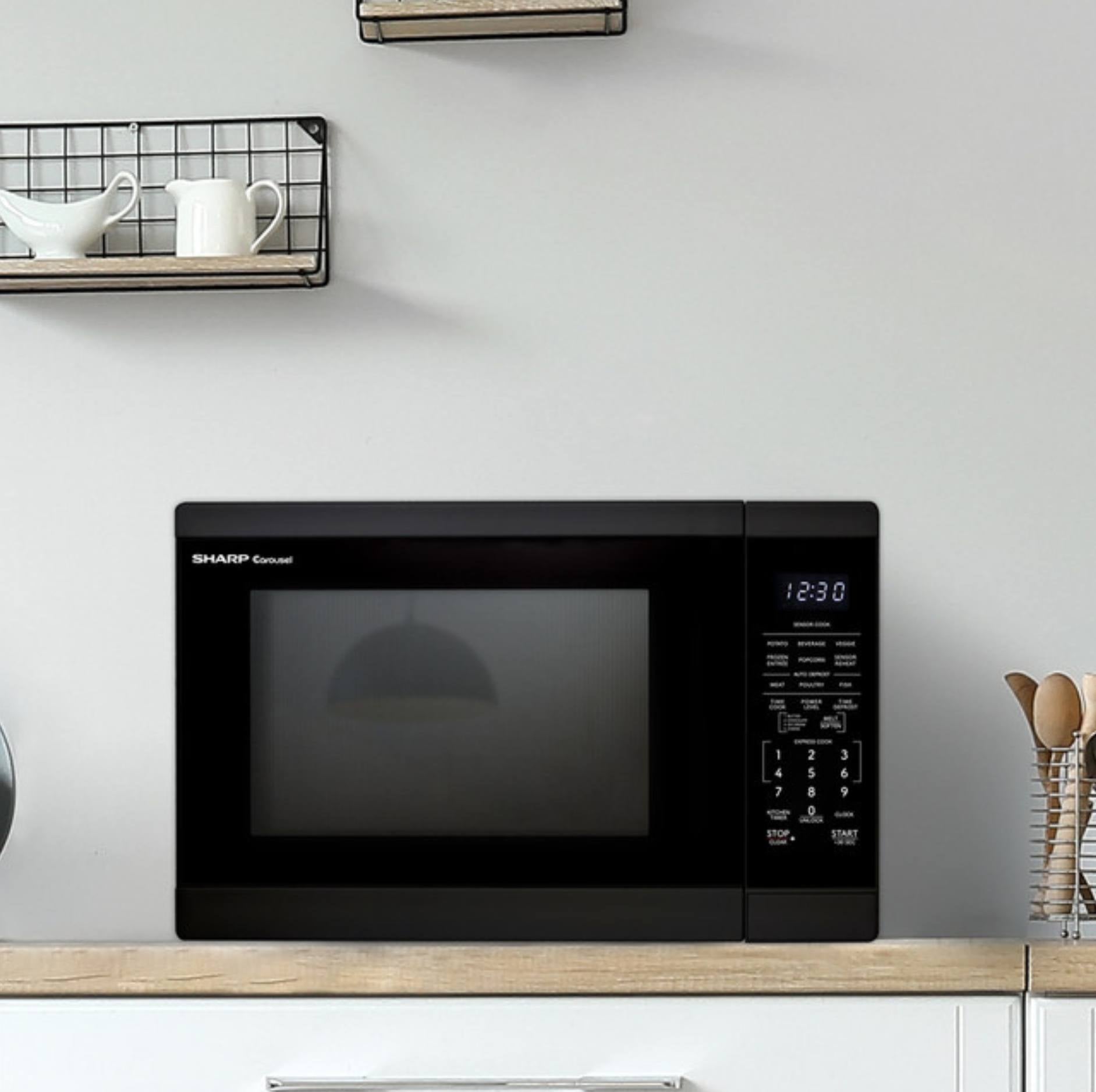 A black Sharp Microwave oven sitting on a light wood countertop in a kitchen setting with a white wall behind it.